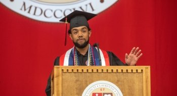 Brian Uceta, wearing academic regalia, stands at a podium and addresses the crowd during Commencement 2023. A scarlet banner with the Saint Lawrence crest is in the background.