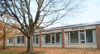 The exterior of Brown Hall on Saint Lawrence University's campus. A large tree stands in front of the building with piles of orange and yellow leaves laying on the ground.