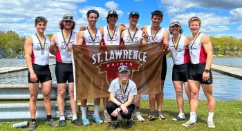 Members of the Saints Men's Rowing Team, wearing white and scarlet uniforms, wear medals around their necks and holding up a brown Saint Lawrence Saints flag, grin as they stand in front of a body of water and celebrate their victory.