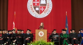 President Kathryn Morris, wearing Commencement regalia, stands behind a podium and addresses the crowd at Commencement. A large scarlet and brown banner with the Saint Lawrence crest hangs in the background.