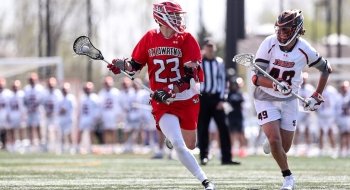 Chris Jordan, wearing a scarlet lacrosse jersey, cradles the ball away from the competition on the lacrosse field. A large group of athletes, wearing white jerseys, stand out of focus in the background.