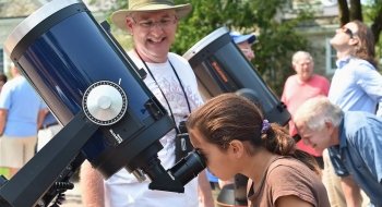 A child with brown hair in a ponytail looks into a telescope while a professor in a white t-shirt and a bucket hat looks on.