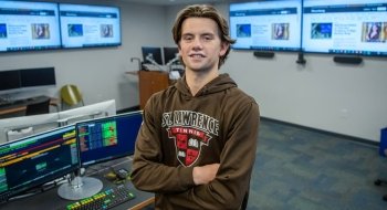 John Hill-Edgar, wearing a brown Saints Tennis sweatshirt, stands in the Bloomberg terminal with his arms crossed. There are four large televisions and two computer monitors in the background.