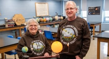 Aileen O'Donoghue and Jeffrey Miller wear brown Saint Lawrence University Gateway to the Path of Totality sweatshirts and stand in a physics lab.