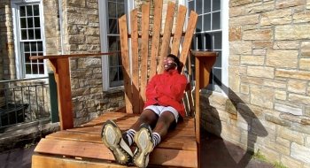A person, wearing a scarlet jacket and eclipse sunglasses, sits on a giant Adirondack chair and smiles up at the sun.