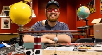 Tyler Karasinksi, wearing red solar eclipse glasses, crosses his arms on top of a desk that is covered in eclipse memorabilia and model of the solar system.