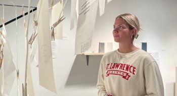 Photo of Saint Lawrence Female Student wearing a white Saint Lawrence University Crew Neck, staring intently at the three dimensional art piece pertruding from the wall. The photo is taken through the art of her. The artwork is a closeline style display of papers with words hanging.