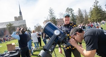 A student peers into a giant black telescope while astronomer Jeff Miller, wearing a black and gold celestial shirt, grins. Gunnison Memorial chapel is in the background.
