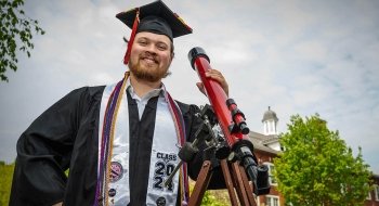 Tyler Karaskinski, dressed in full graduation regalia, stands next to a red telescope in front of a brick building. It's a cloudy day with some sun peeking through.