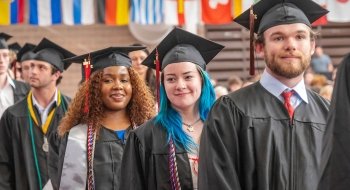 A group of diverse St. Lawrence graduates wearing black graduation caps and gowns stand in a line, smiling. The graduates include a woman with curly brown hair, a woman with blue hair, and a man with a beard. There are nternational flags in the background. The mood is celebratory and proud.