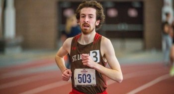 A young male athlete wearing a brown and red St. Lawrence University track uniform with the number 503, running on an indoor track with a determined expression. The background is blurred, highlighting his focus and motion.