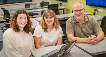 From left to right, Norah Kuduk and Amanda Gagne sit with Professor Ed Harcourt at a desk in a classroom setting with a laptop in front of them. The classroom has chairs and tables with computers in the background.