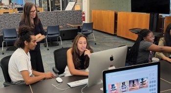 A group of four people are gathered around a large computer in a modern workspace. Two individuals are seated at the computer, focused on the screen, while another person stands behind them, smiling and observing. A fourth person points at the screen, contributing to the discussion. The workspace has a collaborative atmosphere with a mix of seating, workstations, and an open layout. The computer screens display cartoonish images of spiders wearing top hats, indicating a creative project or discussion.