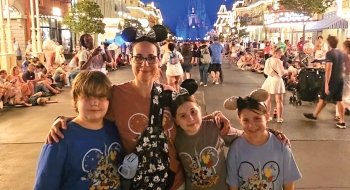 Ronnie Olesker stands with her three kids at Disney. They are wearing Mickey Mouse ears and themed t-shirts. The park is lively, with many visitors in the background, and a brightly lit castle is visible in the distance. The atmosphere is festive and joyful.
