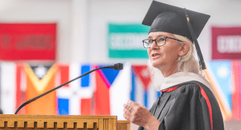 A person in a graduation cap and gown stands at a wooden podium, speaking into a microphone. The individual, who appears to be a faculty member or guest speaker, has white hair and wears glasses. In the background, colorful flags representing various countries or institutions are slightly blurred, creating a vibrant backdrop for the scene. The speaker's expression is focused and engaging, emphasizing the importance of the occasion.