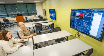 Ed Harcourt and Norah Kuduk sit in a classroom and focus on a large wall-mounted screen displaying a coding interface and a mobile app prototype.