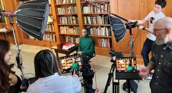 A behind-the-scenes look at an AI video recording session in a library. A person sits in front of bookshelves, facing the camera, while a crew sets up lighting and sound equipment. Several people monitor the recording on screens, ensuring the setup is correct. The atmosphere is professional and focused, with attention to detail in the filming process.