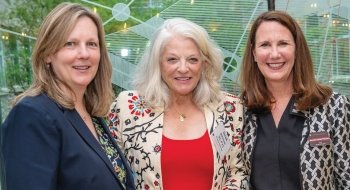 President Morris stands on the left, smiling warmly, wearing a dark blazer and a patterned blouse. In the center is Sarah Johnson, wearing a floral-patterned jacket over a red top, with white hair styled loosely around her shoulders. On the right is Jennifer Curley Reichert, also smiling, wearing a patterned jacket over a black blouse. The three women are close together, appearing at a formal event, with a modern glass backdrop featuring geometric designs behind them. The atmosphere is professional yet frie