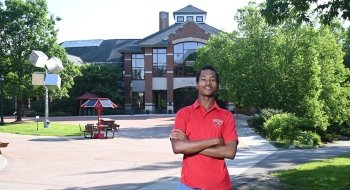 Joshua-Daniel Alleyne, wearing a scarlet polo t-shirt, stands in front of the Sullivan Student Center on Saint Lawrence University's campus.
