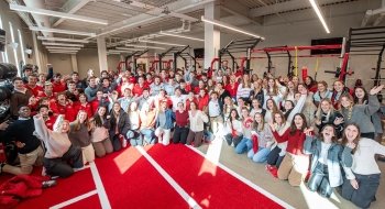 A large group of Saint Lawrence University students, many dressed in red and gray, gather excitedly in the campus fitness center, posing and cheering. They stand in front of workout equipment under bright overhead lights. The group is smiling and waving, creating a lively and celebratory atmosphere.