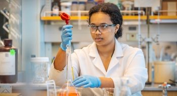 A person wearing safety goggles, gloves, and a lab coat carefully uses a pipette in a laboratory setting. Shelves filled with bottles and equipment are visible in the background. The person appears focused and precise in handling the laboratory tools.