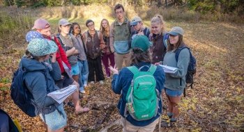 A group of students gathers in a circle outdoors, listening attentively to an instructor with a green backpack. The group is standing on a leaf-covered forest floor, holding notebooks and dressed for a field experience. Sunlight filters through the trees, creating a warm, autumn atmosphere.