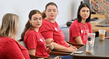 Four students sit at a table, engaged in a discussion. They are wearing matching red Saint Lawrence University shirts, and several iced coffee drinks are on the table. The background shows a bright classroom with a colorful abstract design on the wall.