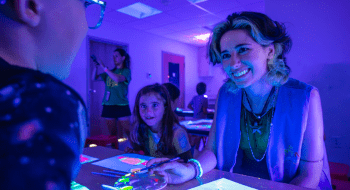 Alexandria Nye sits at a table with young children as they work on a glow in the dark project while black lights shine on them. Everyone is illuminated in purple light.