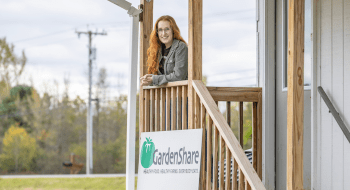 Sonja Jensen  leans on the wooden railing of a porch, smiling. A &quot;GardenShare&quot; sign is visible below, featuring the slogan &quot;Healthy food. Healthy farms. Everybody eats.&quot; The background includes trees and power lines on a cloudy day.