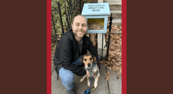 Sean Kelly crouches on a sidewalk, smiling while holding a dog on a leash. Next to them is a small blue box labeled &quot;Stevie's Biscuit Bar,&quot; filled with dog treats. Fallen leaves and a black metal fence frame the scene.