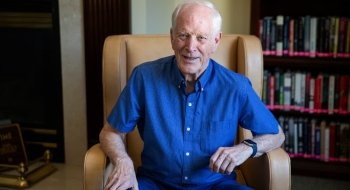 Thomas Wallace, wearing a blue button up t-shirt, sits in a leather chair in a library.