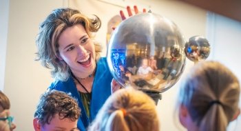 A smiling instructor in a blue jacket shows a group of children a reflective metal sphere during a science demonstration. The children look on curiously, and their reflections are visible in the shiny surface of the sphere.