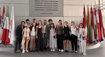 A group of individuals stands in front of the European Central Bank wall, with various national flags displayed on either side. They are smiling and dressed in a mix of casual and formal attire. The setting suggests a visit or event at the bank.