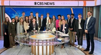 A group of students and faculty from Saint Lawrence University pose together in the CBS News studio. They stand behind a glass desk with the CBS News logo displayed prominently in the background. Everyone is smiling, dressed in professional attire, and wearing ID badges.