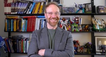 Dan Look, wearing a blazer and a green t-shirt, smiles and stands in front of shelves filled with transformer action figures.