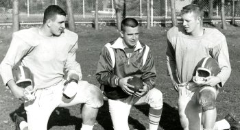 Three football players kneel on a grassy field, two wearing practice uniforms and holding helmets, while the one in the center wears a jacket and holds a football. A chain-link fence and trees are visible in the background. The image appears to be from a vintage setting, showcasing a practice or team discussion.