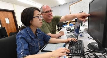 Ed Harcourt and a Saint Lawrence student sit in front of a computer in a lab and collaborate on a project. Harcourt points at something on the computer screen as the student's hands hover over the keyboard.