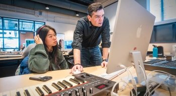 Chris Watts, leans on a desk and peers into a computer monitor. A student, wearing a headset, observes. There is a large electronic keyboard next to the computer.