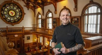 Howard Eissenstat, wearing a black turtle neck, holds a book and stands inside the Herring Cole building on Saint Lawrence University's campus. There is a large, circular, stained glass window in the background.