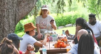 Sara Ashpole, sits on a picnic table outside and holds a jar of jam. She is speaking to a group of students gathered around picnic tables. There are fresh vegetables scatter through out the table.