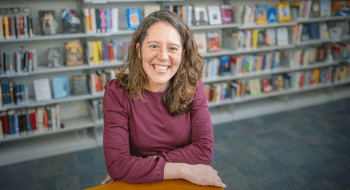 Sarah Barber, wearing a maroon long sleeve shirt, leans on a bookshelf in a library.  In the background, large bookshelves are filled with vibrant colored books.