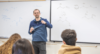 Sam Tartakoff teaches chemistry, gesturing while explaining concepts on a whiteboard filled with chemical structures. Students sit in the foreground, listening attentively.