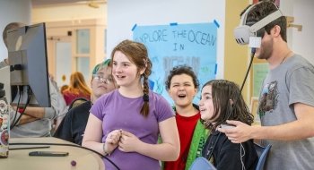 A group of kids excitedly watch as a person uses a virtual reality headset in a classroom setting. A sign in the background reads “Explore the Ocean.” One child holds a controller while others smile and laugh.
