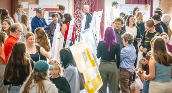 A large group of students, faculty, and visitors gather around poster displays at an indoor academic event, engaging in discussions and viewing presentations.