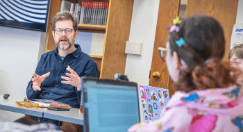 Adam Harr speaks during a small classroom discussion, gesturing with both hands. A student in the foreground listens while using a laptop decorated with colorful stickers.