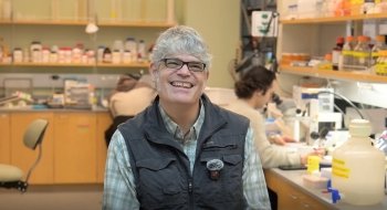 Alex Schreiber smiles while seated in a science lab. Lab equipment, bottles, and another person using a microscope appear in the background.