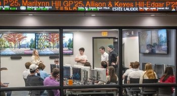 A professor speaks to a group of students seated at computer stations in a modern classroom at Saint Lawrence University. Large monitors display a fall campus scene, and a digital stock ticker scrolls above the glass wall.