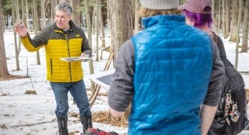 Jon Rosales leads a discussion in a snowy forest at Saint Lawrence University, holding a clipboard while gesturing toward students in winter gear. The group stands among trees with snow-covered ground, participating in an outdoor learning session.