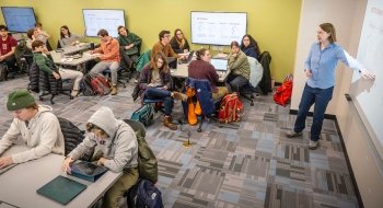 Lisa Torrey stands at a whiteboard pointing to a diagram while students sit at tables in small groups, listening, writing, or working on laptops in a bright, modern classroom.