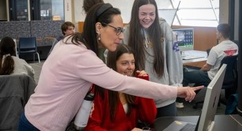 A faculty member at Saint Lawrence University points at a computer screen while two students smile and look on. They are gathered around a table in a bright, open study area with other students working in the background.